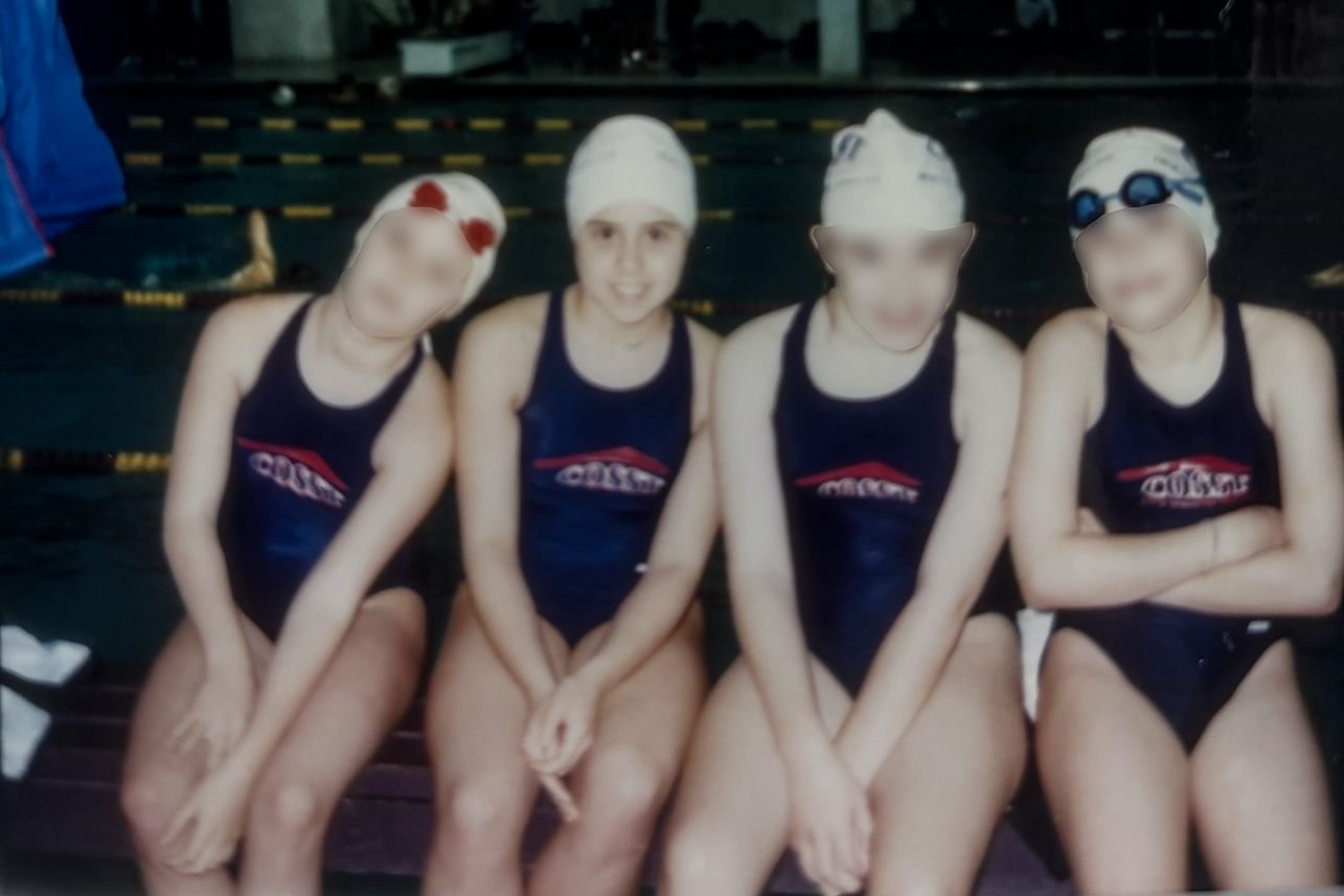 Four young swimmers wearing swimsuits and caps sit together on a bench at an indoor pool