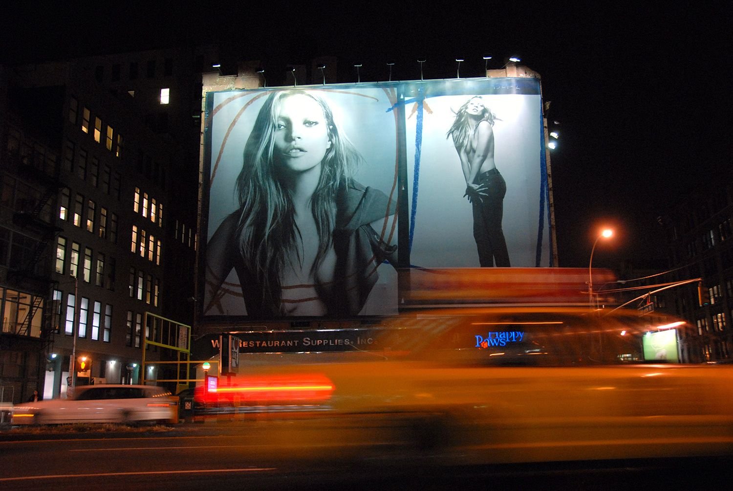 A night-time city street shows a large black-and-white fashion billboard above blurred taxi lights