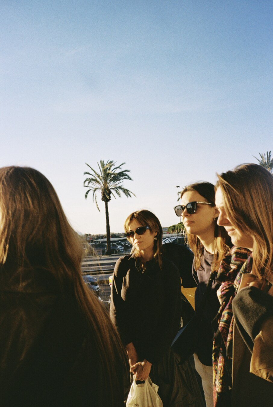 Four women wearing sunglasses stand together outdoors with a palm tree and parking lot visible behind them under a clear blue sky