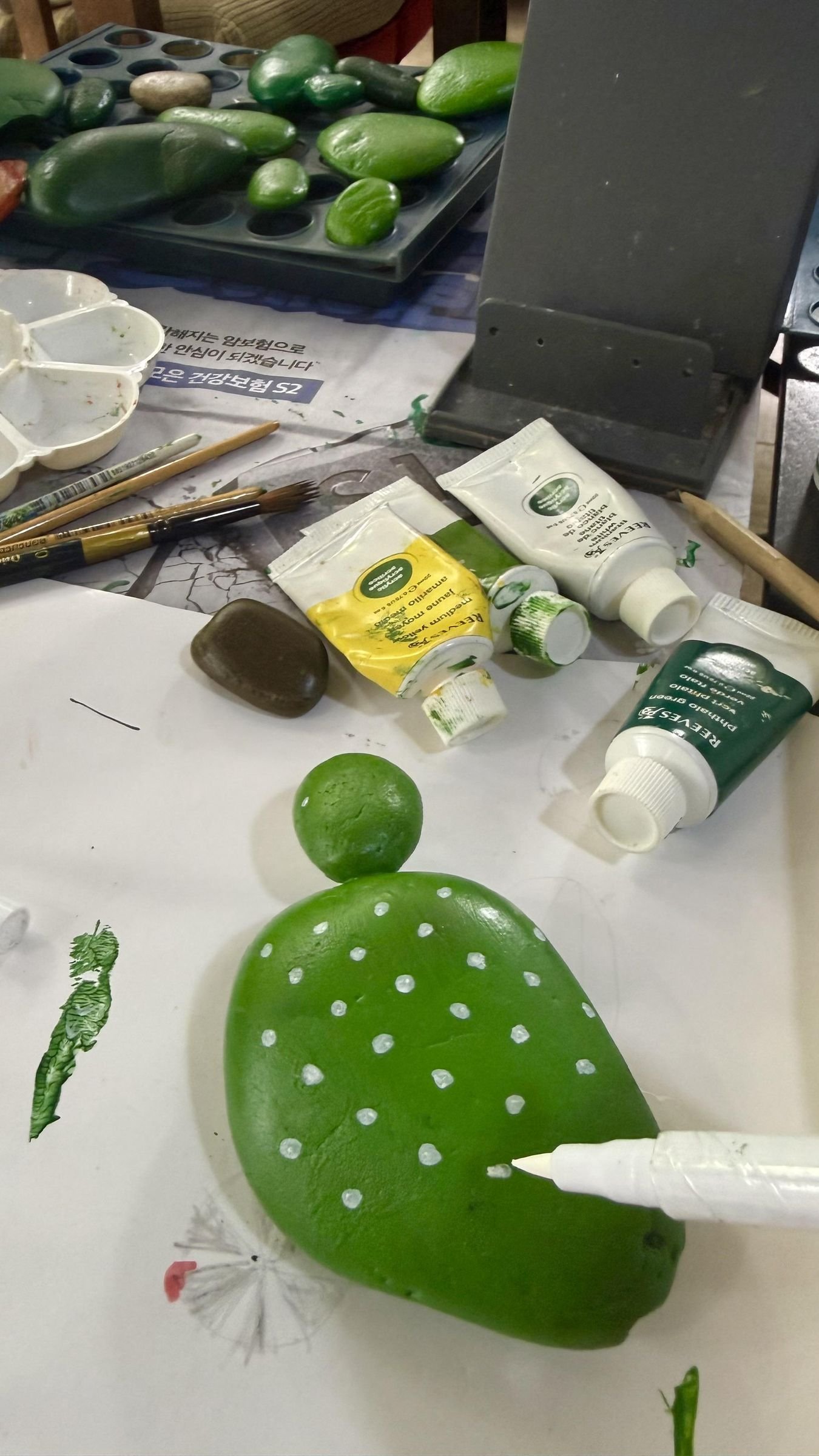 A green-painted cactus stone on a craft table beside acrylic paints and brushes