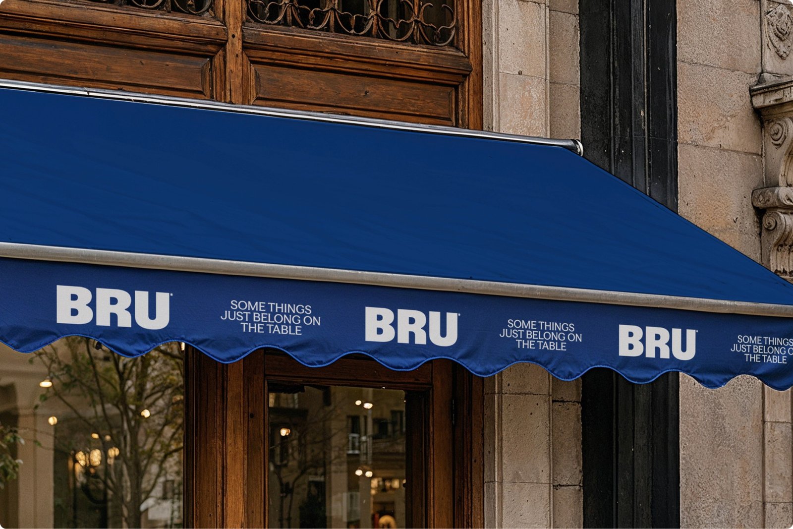Blue awning above a restaurant entrance featuring BRU branding and "Some things just belong on the table" tagline, with elegant wooden doors and stone facade behind