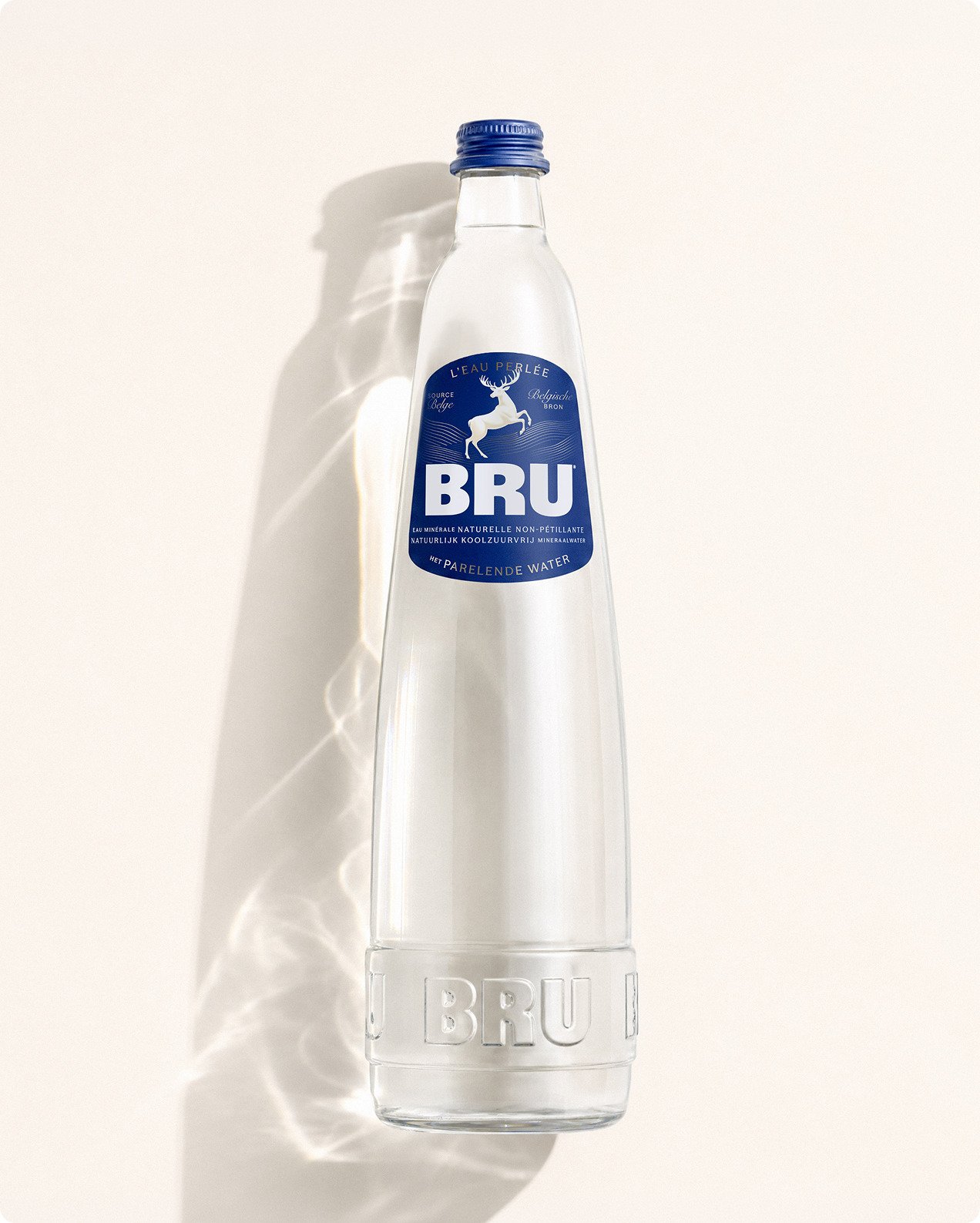 Clear glass bottle of BRU sparkling water with blue label featuring a deer motif and blue screw cap, casting a soft shadow against a cream background