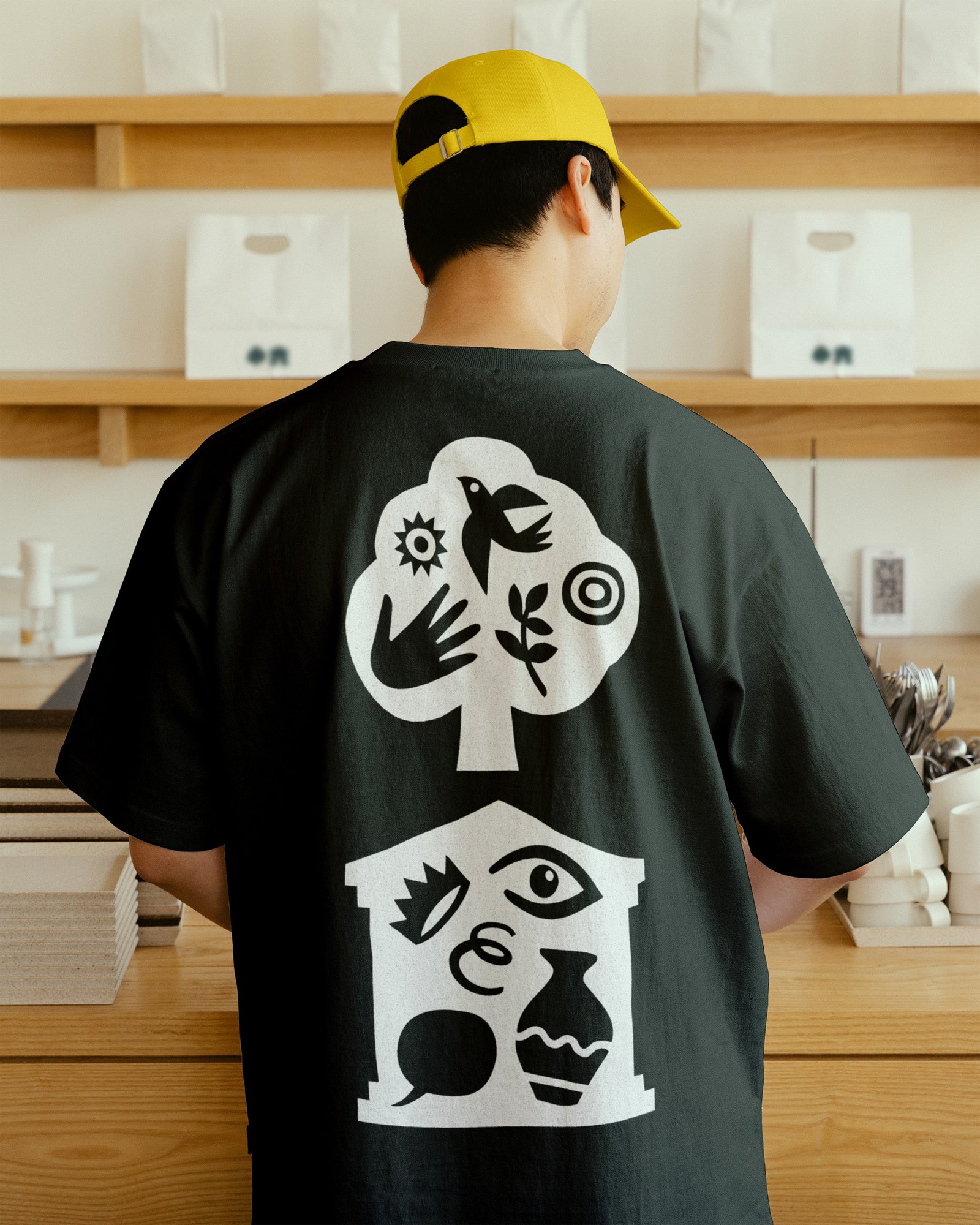 Person wearing a yellow cap and black T-shirt printed with white abstract symbols representing a tree, house and natural elements from Gunnersbury's rebrand