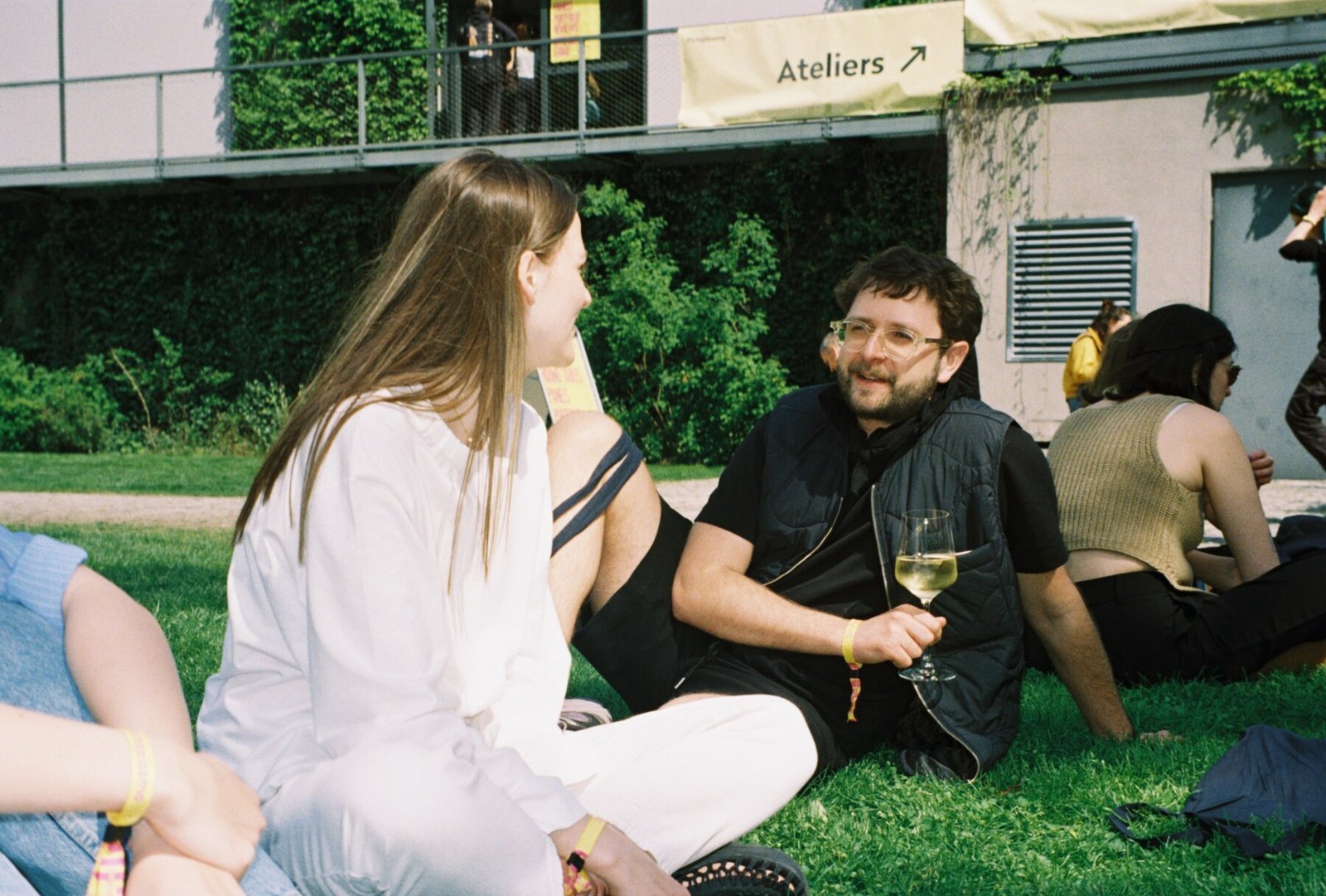 People socialise and drink wine outdoors on grass at an andstudio Wine Design Exchange event, with ivy-covered building and Ateliers sign visible behind them