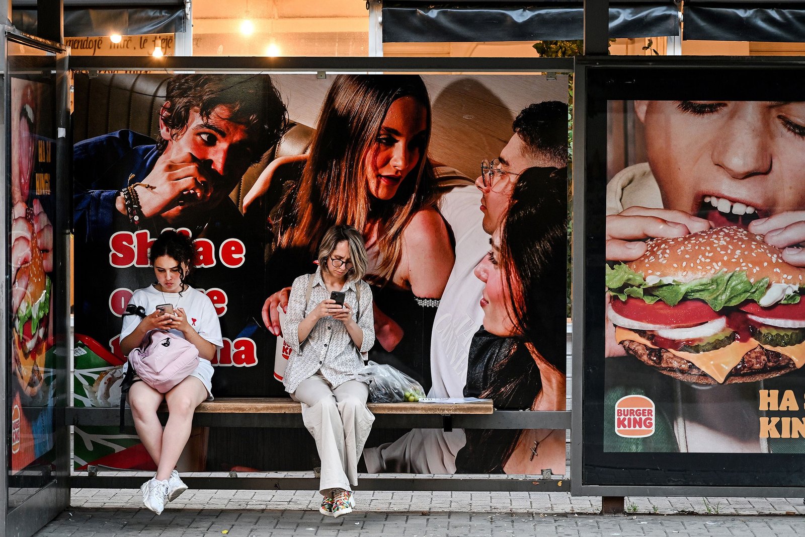 Two women sit at a bus stop using their phones beside large fast-food advertisements