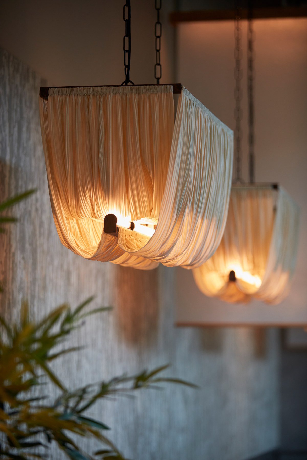 Hanging pendant lights with cream-coloured pleated fabric shades suspended from black chains, glowing warmly against white walls with potted plants nearby