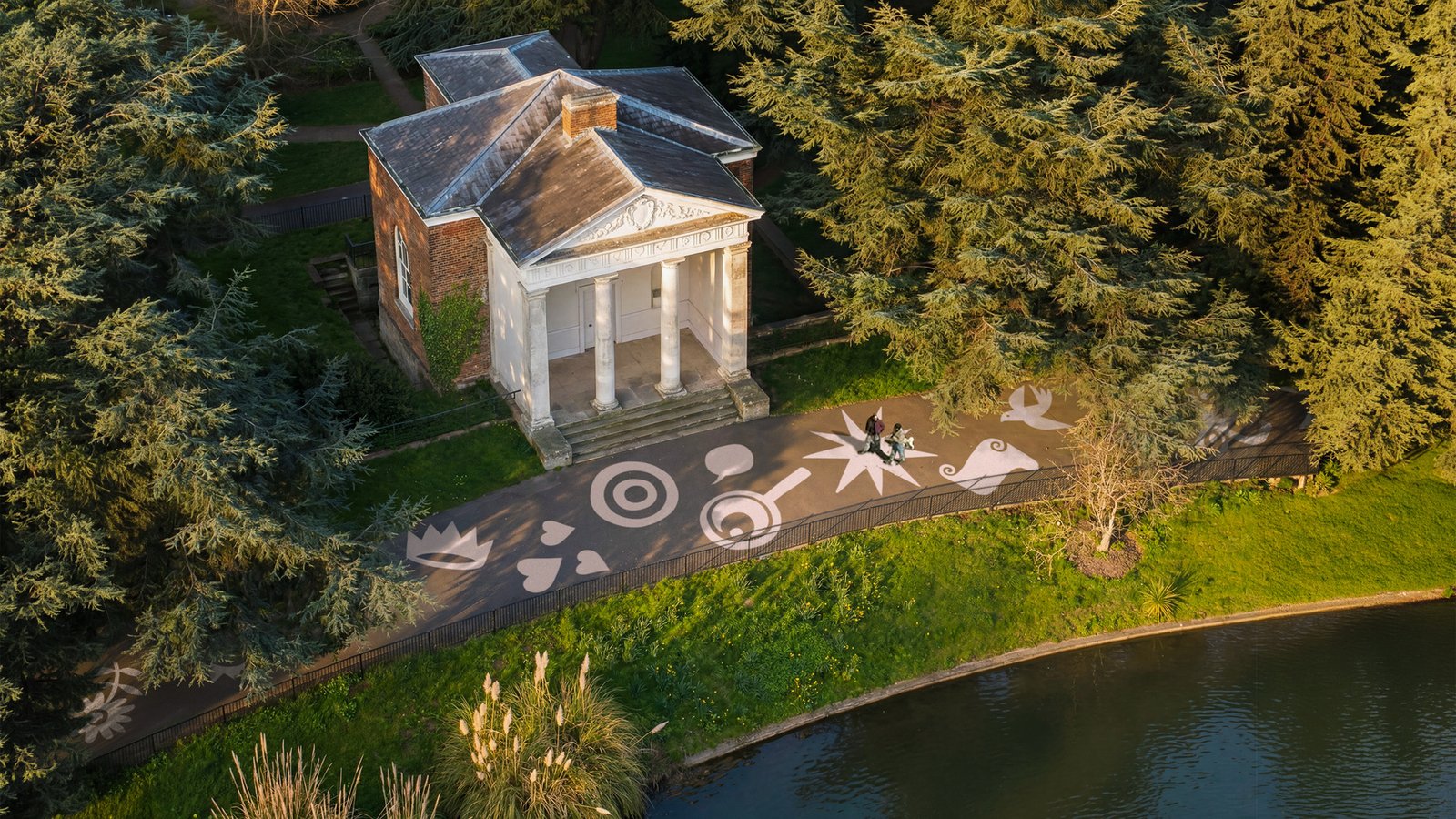 Aerial view of Gunnersbury Museum's classical pavilion surrounded by woodland, with a patterned ground design and boating lake nearby