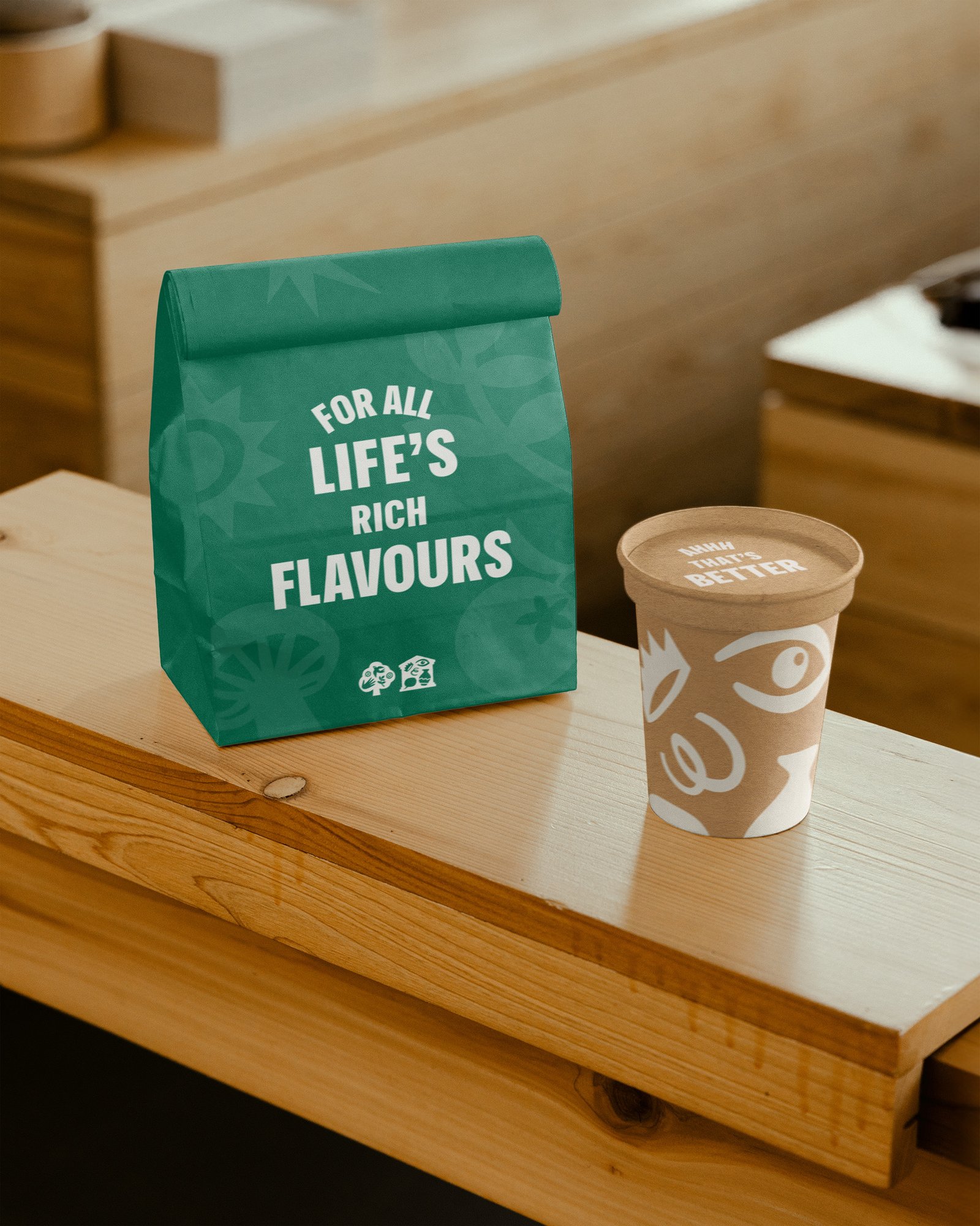 Green paper bag printed with ‘For all life's rich flavours’ next to a brown cup with white patterned design on a wooden counter