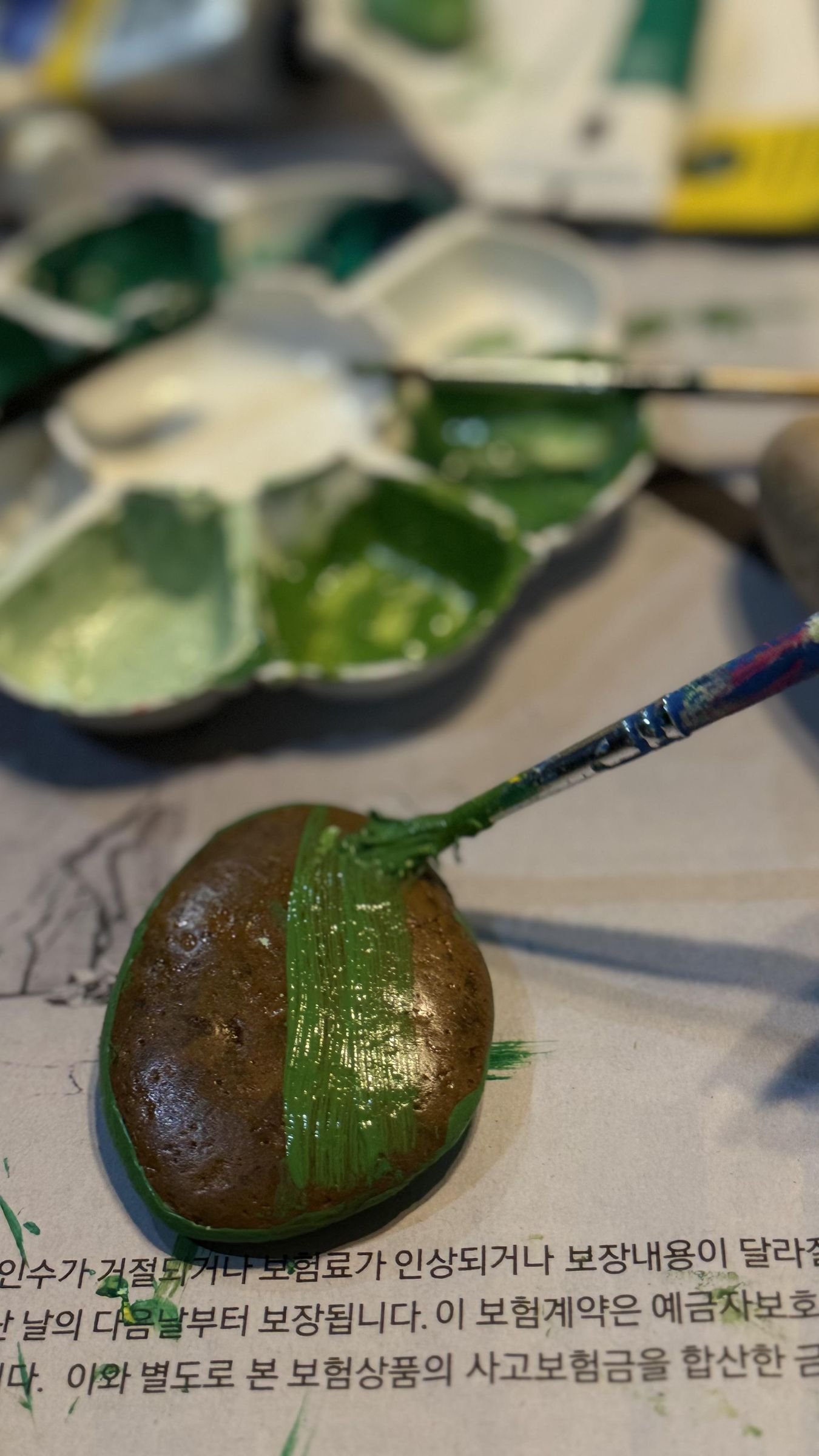 A paintbrush applying the first green acrylic coat to a rounded stone