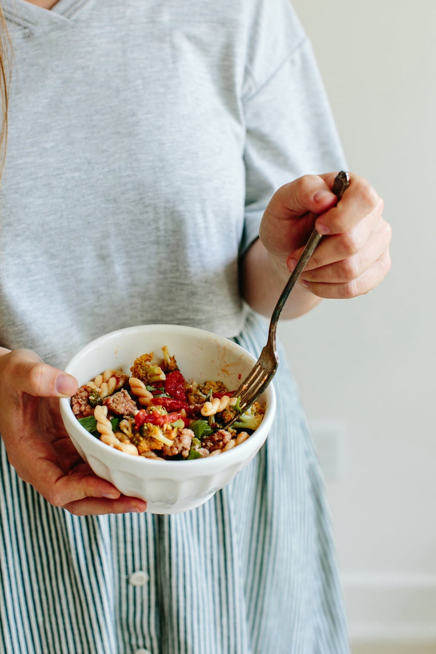 A person holding a bowl of gemelli with turkey sausage and broccoli