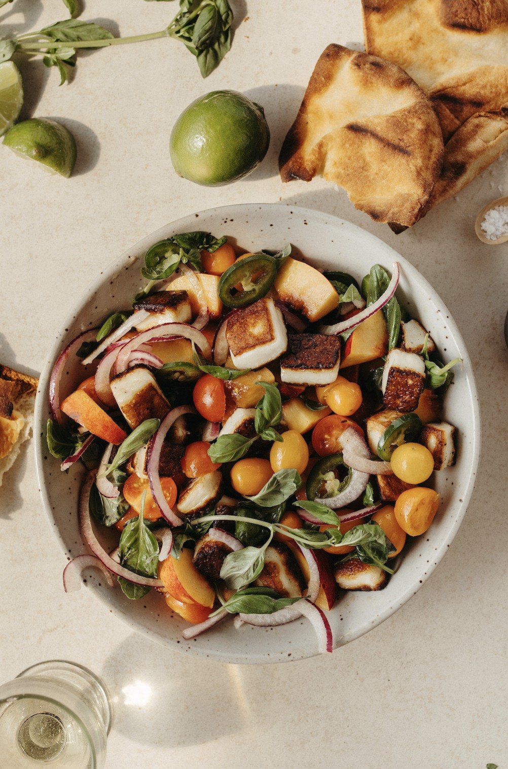 Overhead tomato, nectarine, and halloumi salad beside grilled pita and limes