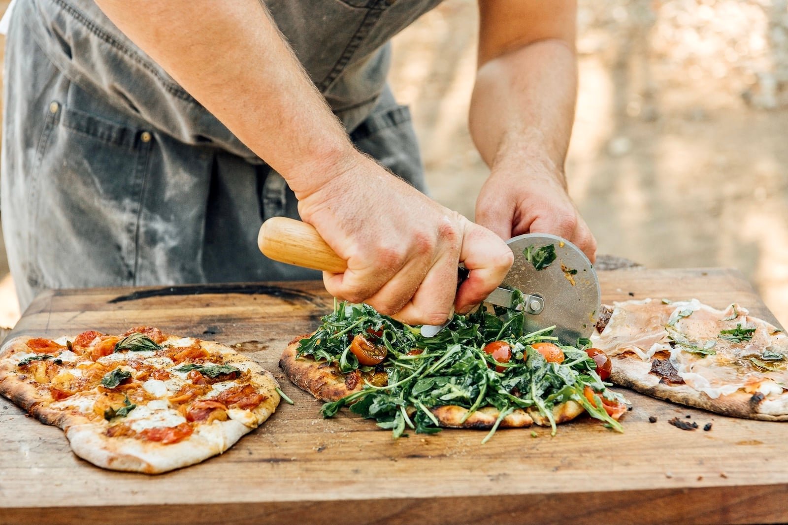 Three pizzas being sliced on an outdoor table