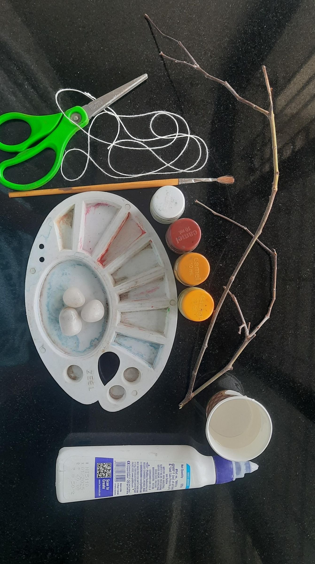 Pebbles, paint, brushes, glue, string, scissors, and a twig arranged for the craft