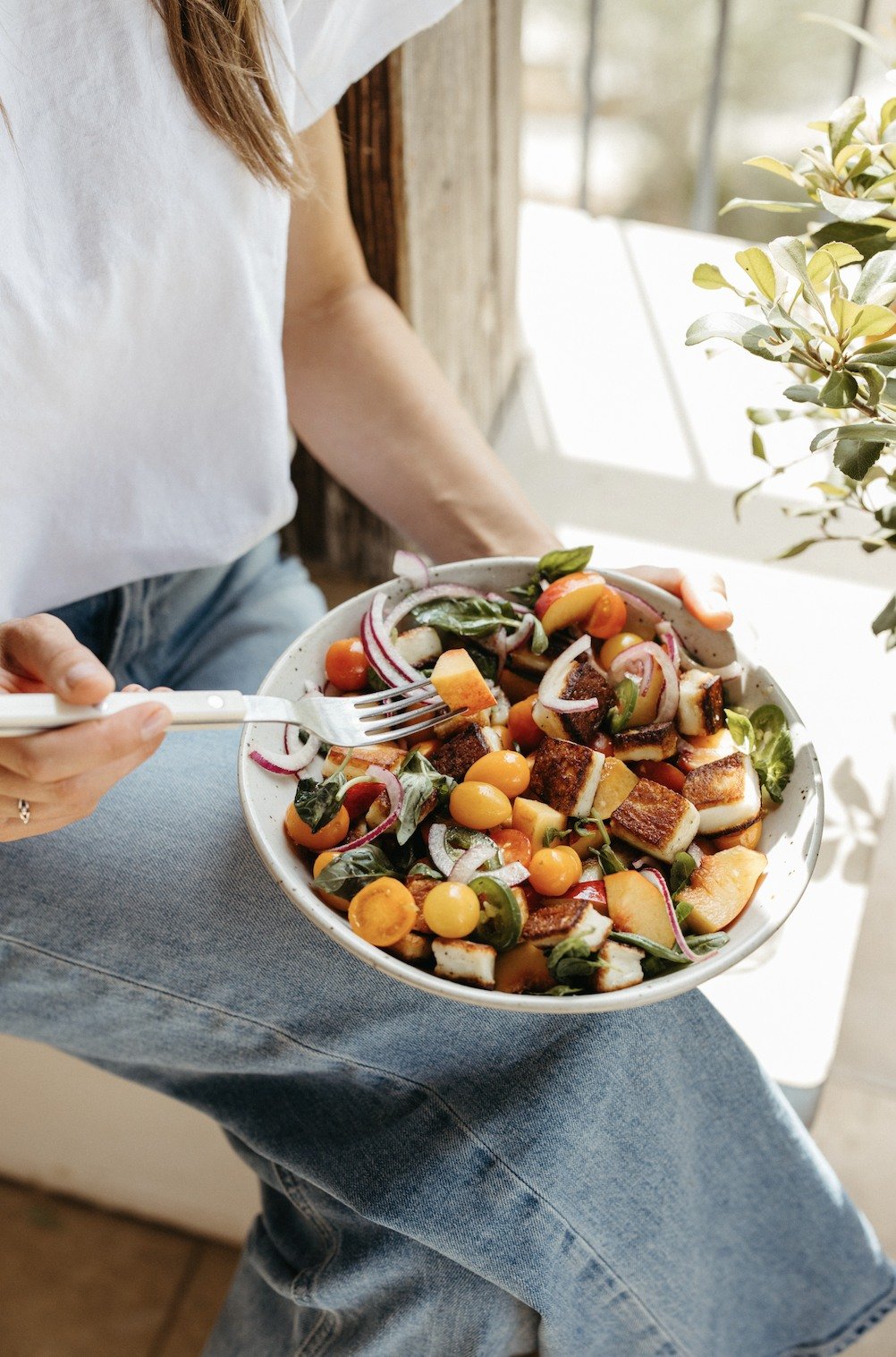 A woman holding a bowl of tomato, nectarine, and halloumi salad on her lap
