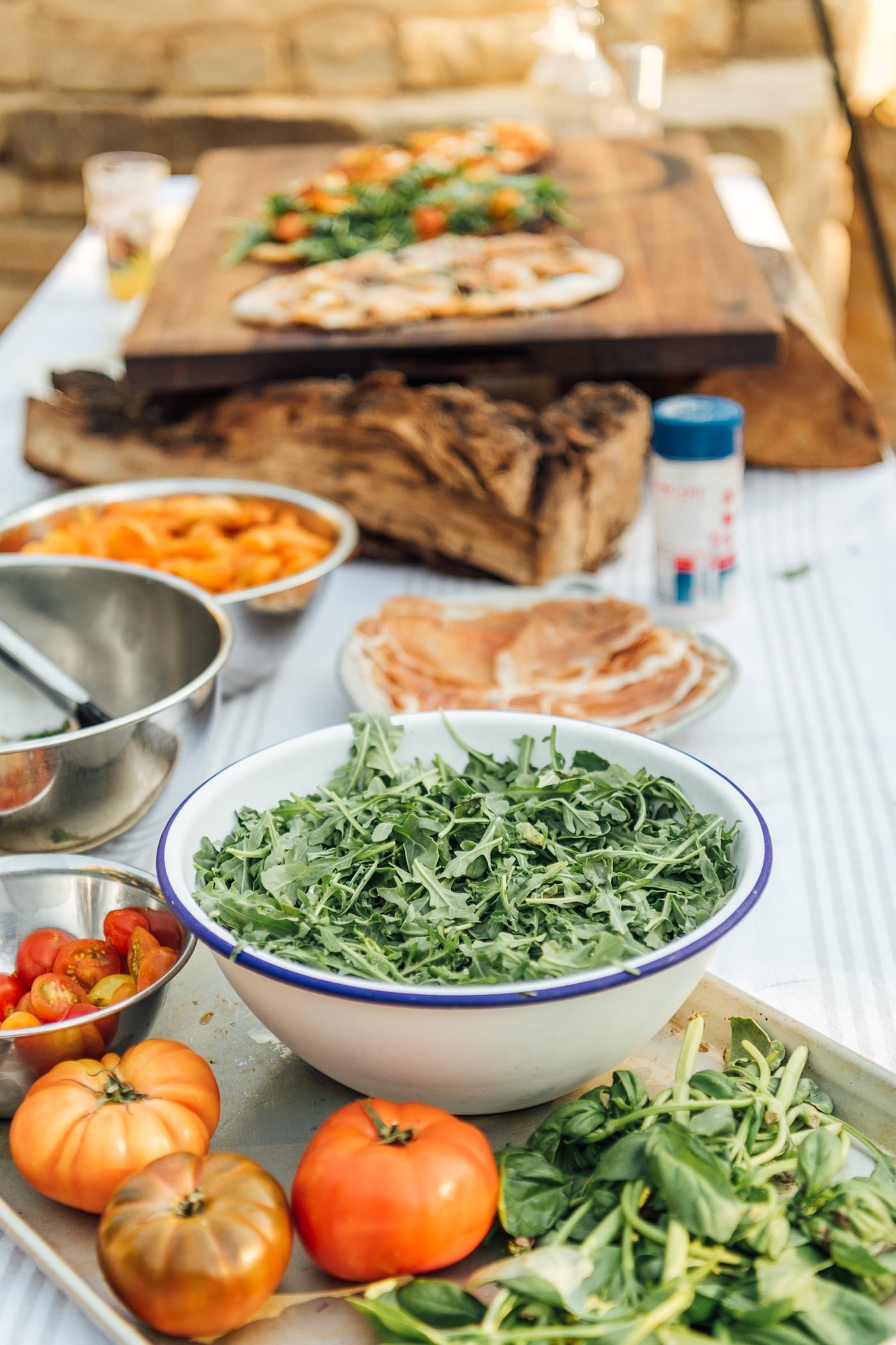 An organized outdoor pizza topping bar with bowls of greens and tomatoes